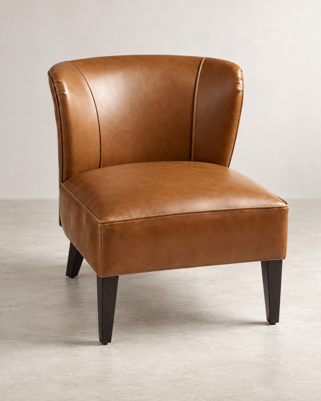 Warm cognac upholstery follows a softly curved back and generous seat. Subtle piping and dark tapered legs give this compact chair a tailored finish.