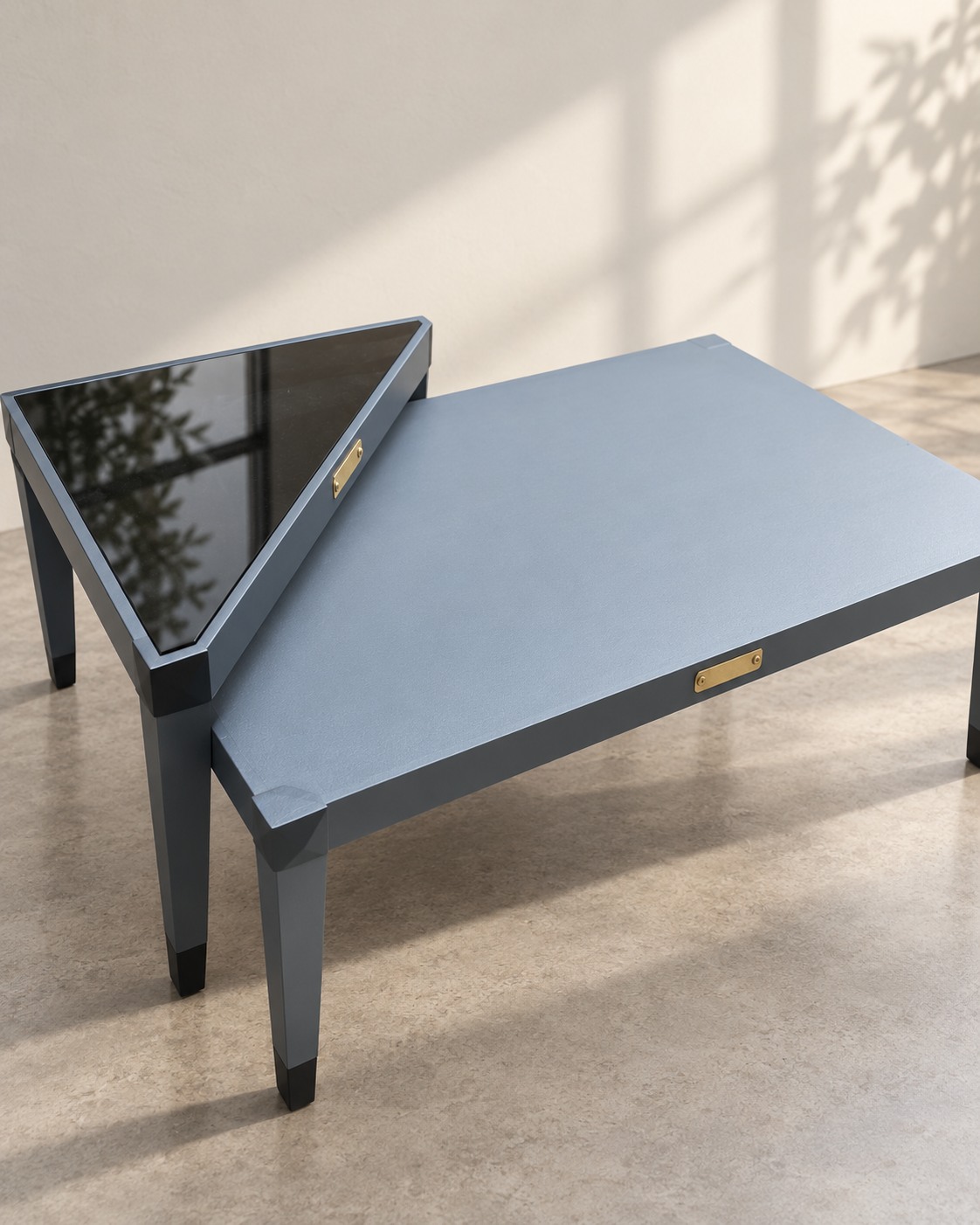 Angular forms pair a muted slate surface with a dark reflective tabletop. Small gold-toned details and contrasting feet add definition to this geometric table arrangement.