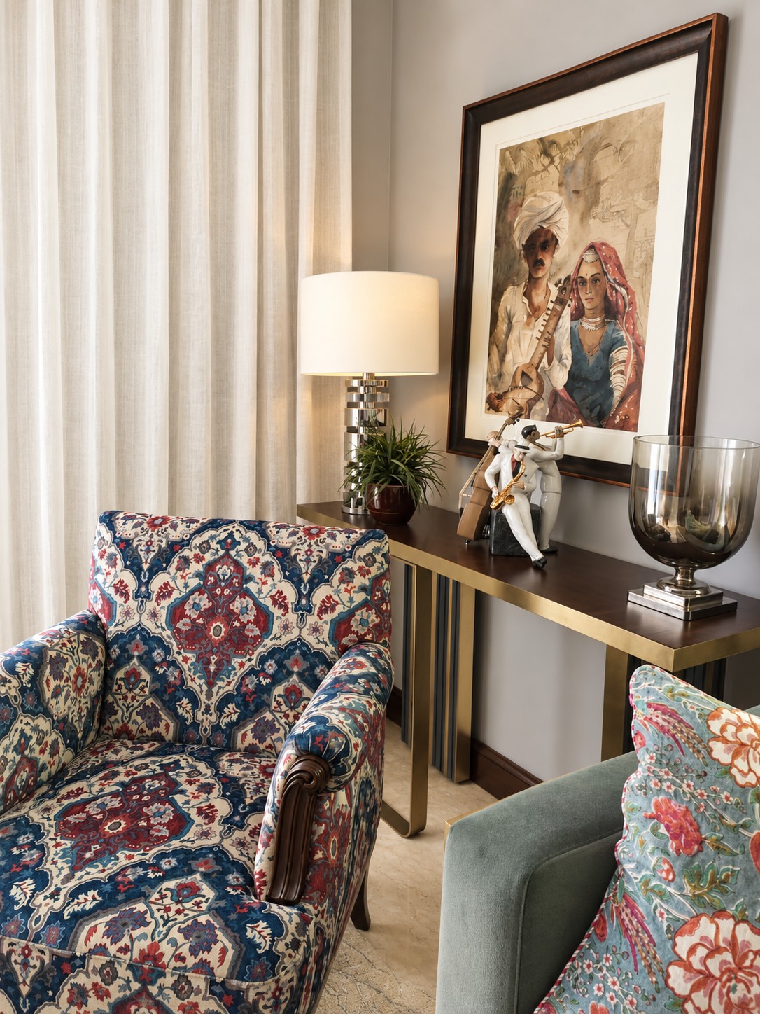 Patterned armchair beside a console with framed figurative art, a lamp, greenery and decorative objects.